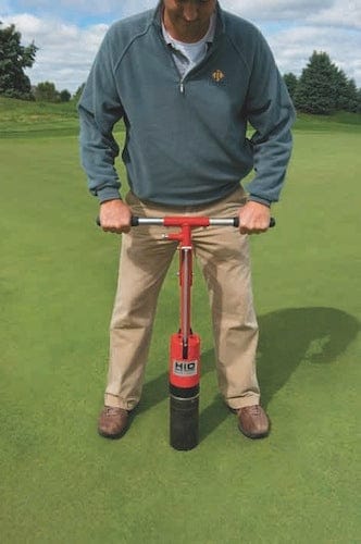 Lochbohrer mit rotem Griff und schwarzem Bohrzylinder im Einsatz auf grünem Golfplatz.